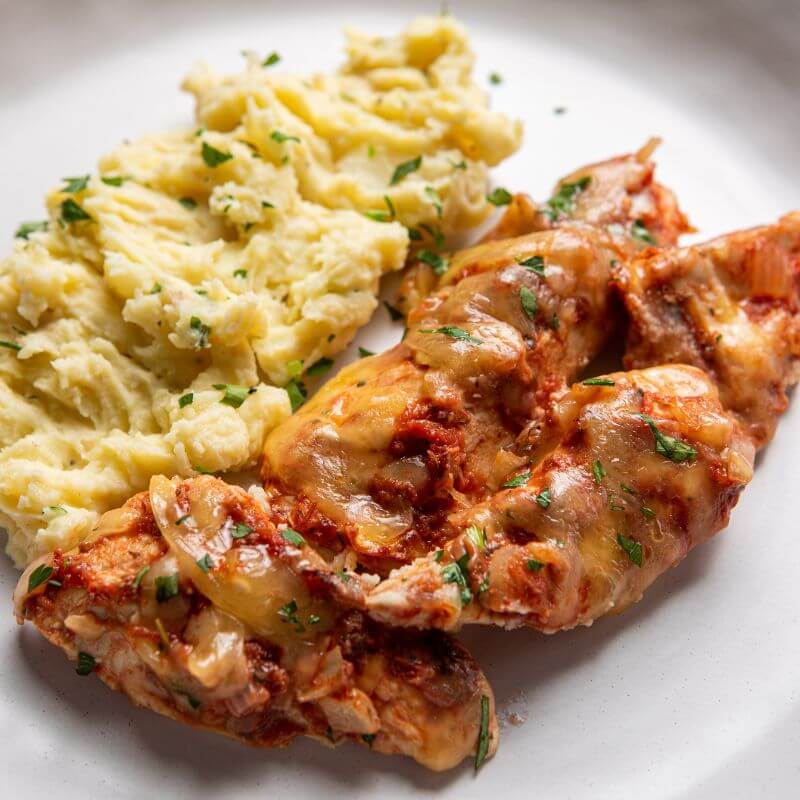 A close up of chicken, covered in a rich tomato sauce and melted cheese, served on a white plate with creamy white mashed potato.