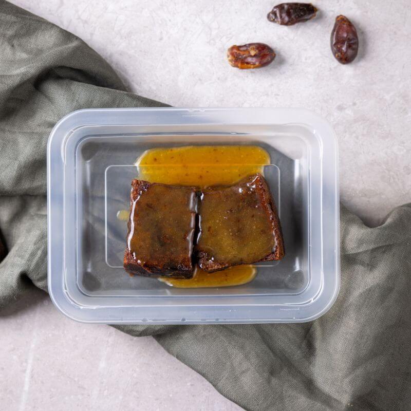 A top-down photograph of two serves of sticky date pudding surround by sauce in a container, served on a green grey cloth on a marble table, with a few dates surrounding the dessert.