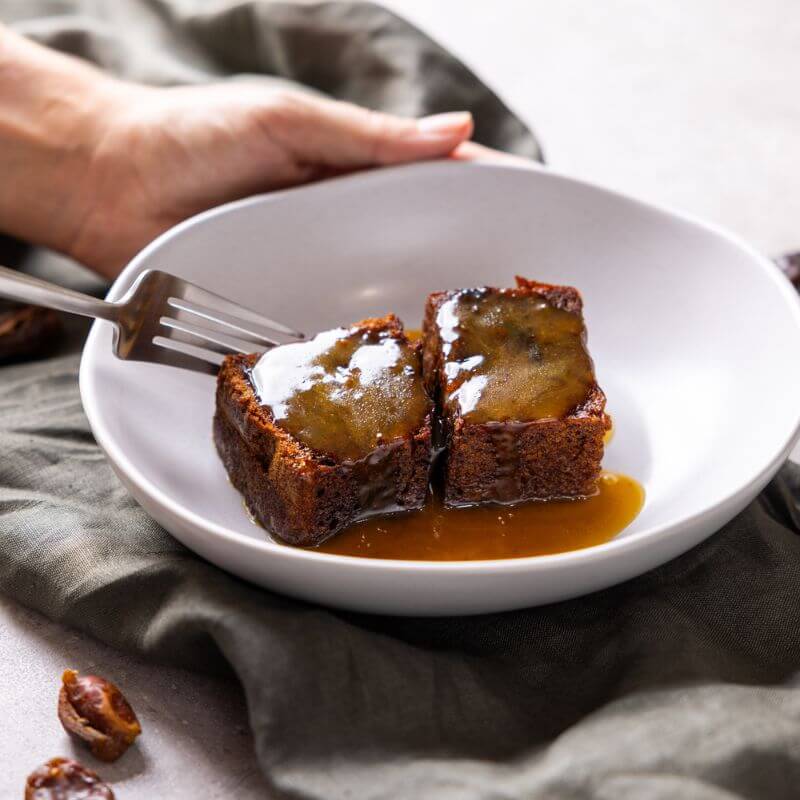 An action photograph of a fork slicing into one of two sticky date pudding servings in a white ceramic bowl covered in a golden sticky date sauce. A hand is holding the bowl, and the bowl is sitting on a green grey cloth on a marble bench.