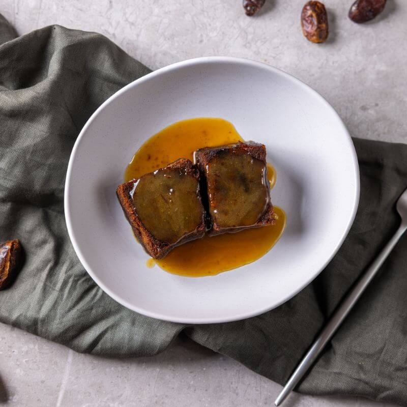 A top-down photograph of two serves of sticky date pudding surround by sauce in a white ceramic bowl. The bowl is sitting on a green grey cloth on a marble bench top, with a fork on the side and some fresh dates scattered around.