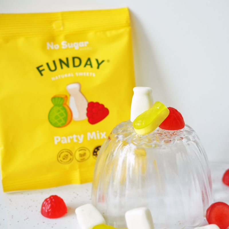 A side on photograph of a small glass dome, with the raspberry, pineapple and milk bottle gummies sitting on top.  The dome is sitting on a marble bench with the packet the gummies are delivered in leaning against a white backdrop behind it. Extra gummy lollies are scattered around the bench.
