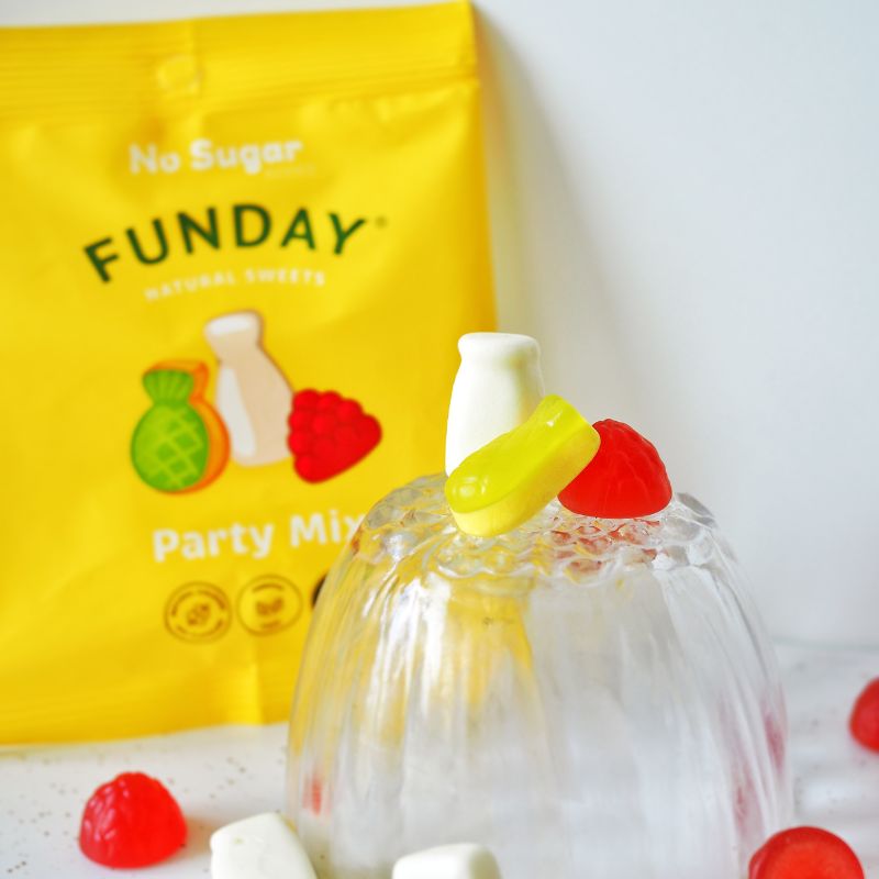 A side on photograph of a small glass bowl containing raspberry, pineapple and milk bottle gummy lollies, served on a white bench top. Behind the bowl is a yellow packet the gummies are delivered in leaning against a white back drop.