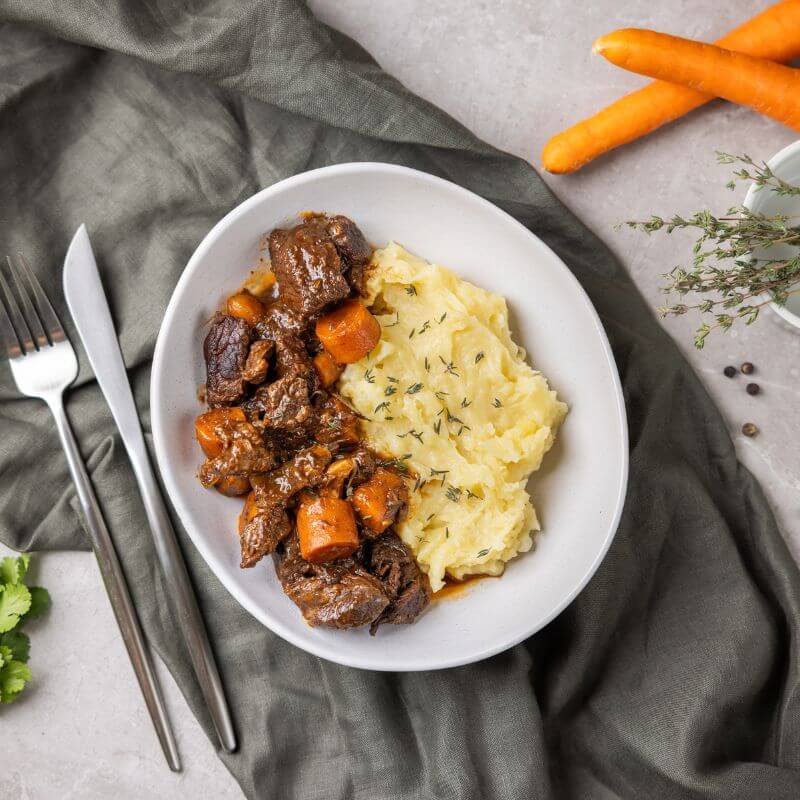 A hearty bowl of Beef Bourguignon sits on a dark olive green cloth, surrounded by fresh carrots, scattered peppercorns, and a garnish of thyme in a white ramekin. A fork and knife rest beside the bowl, adding to the rustic dining scene.
