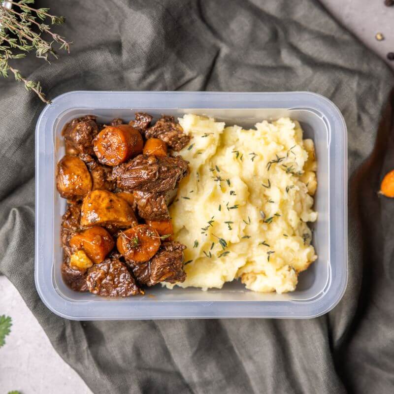 A freshly prepared serving of Beef Bourguignon in a clear container, showcasing tender slow-cooked beef, carrots, and mushrooms in a rich sauce—perfect for a ready-to-enjoy meal.