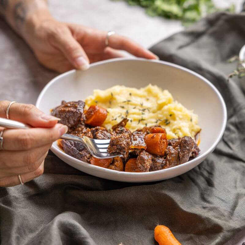A warm and inviting shot of Beef Bourguignon, with one hand holding the bowl and another lifting a tender piece of beef, highlighting its rich texture and slow-cooked goodness.