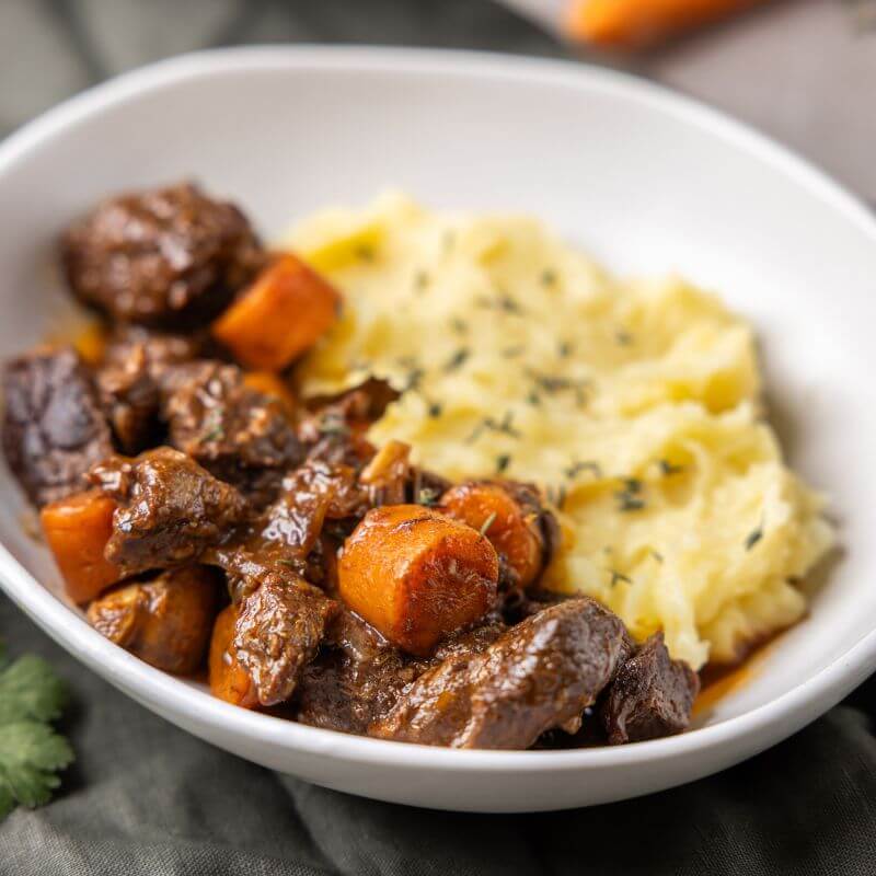A detailed shot of Beef Bourguignon, showcasing tender beef, mushrooms, and carrots coated in a glossy, pomegranate-infused sauce—bursting with rich, slow-cooked flavors.