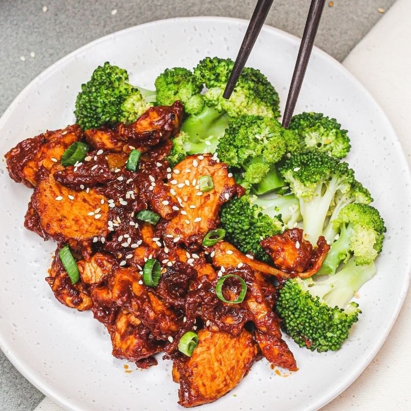 Action shot of chopsticks picking up a piece of broccoli from the Korean Sesame Chicken and Broccoli on the white plate, showcasing the sticky sesame sauce and fresh broccoli.