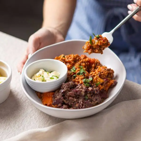 ction shot featuring Chilli Con Carne in a white bowl with sour cream and spring onion, with a person scooping up a portion of the chili using a fork, showing the rich texture and flavours.