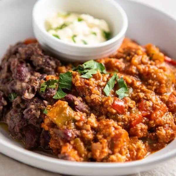 Close-up shot of Chilli Con Carne in a white bowl, highlighting the vibrant chili, sour cream, and spring onion garnish for a mouth-watering view.