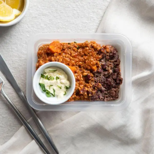Chilli Con Carne in a meal container, with a knife and fork on the side, a garnish of lemon, and a ramekin of sour cream topped with spring onion, capturing the dish’s perfect presentation.