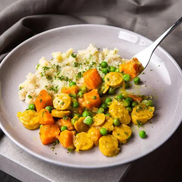 Action shot of curried sausage with cauliflower mash, with a fork digging into the sausage and mash, showcasing the creamy texture of the cauliflower mash and the hearty curried sausage.