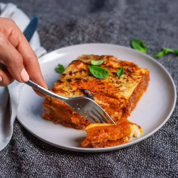 Action shot of a hand holding a fork, cutting into the layers of the Pumpkin Lasagna. This shot captures the creamy, decadent layers of the dish while emphasizing its comforting texture.