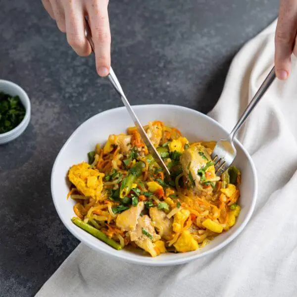 An action shot of Chicken Singapore Noodles with two hands holding a fork and knife, cutting into a piece of chicken. A white ramekin filled with chopped spring onion can be seen in the background