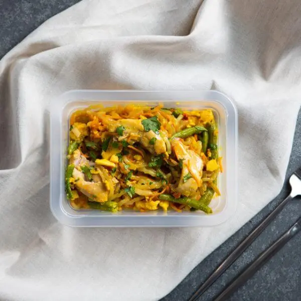 A clear plastic container filled with Chicken Singapore Noodles, featuring tender chicken pieces, vibrant vegetables, and rice noodles. The container rests on a white cloth, with a fork and knife faintly visible in the shot