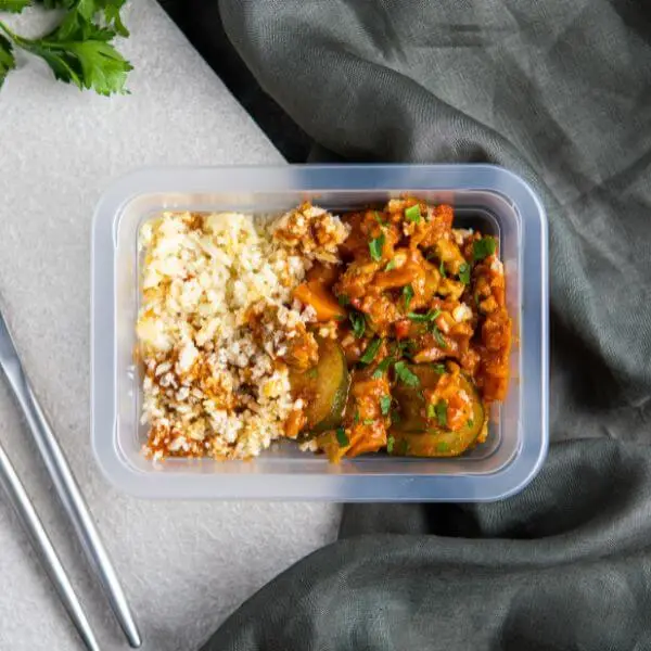  Clear container shot of the Butter Chicken with Cauliflower Rice, perfectly portioned for convenience. The container is set on the grey countertop, and the vibrant garnishes—parsley and the rich orange curry—shine through, emphasizing its freshness and homemade quality.