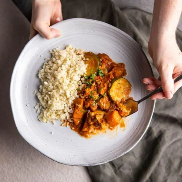 Action shot of one hand holding a plate of Butter Chicken with Cauliflower Rice while the other hand scoops up a piece of chicken. This dynamic image adds a personal and inviting touch, making the dish feel warm and approachable.